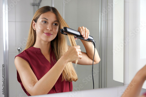 Hispanic young woman using steam straightener to style hair in the mirror on bathroom photo