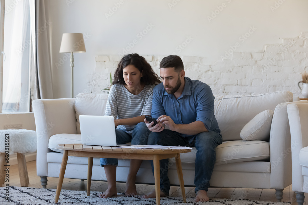 Focused married couple doing paperwork, accounting job at home, using laptop computer, calculator over paper bills, paying insurance, mortgage, rental fees, loan conditions, using online app Focused married couple doing paperwork, accounting job at home, using laptop computer, calculator over paper bills, paying insurance, mortgage, rental fees, loan conditions, using online app