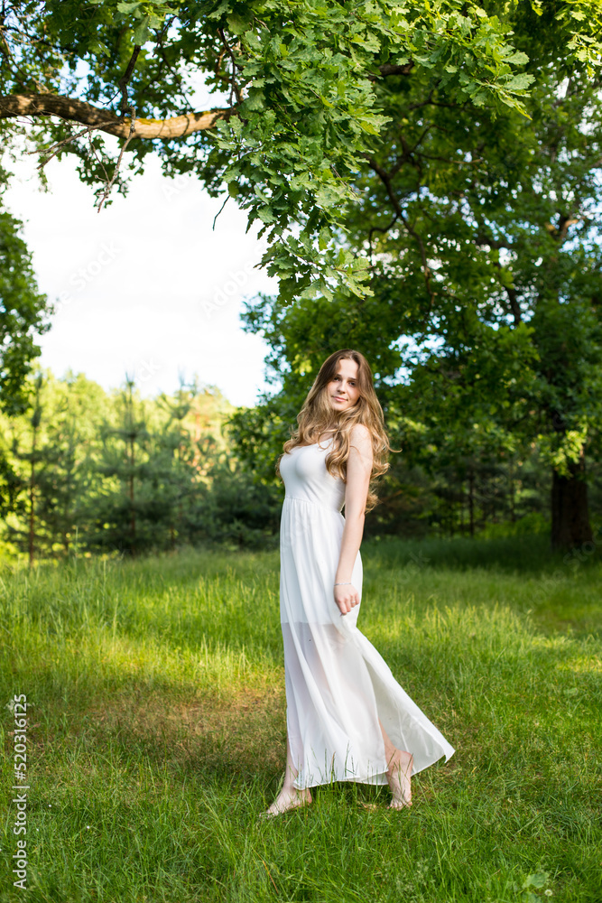 Beautiful young Russian girl dressed in a white dress, walks through the forest among the oaks in the rays of the sunset Beautiful young Russian girl dressed in a white dress, walks through the forest among the oaks in the rays of the sunset