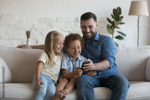 Happy millennial father teaching cheerful little daughter and son kids to use online shipping app on mobile phone, taking selfie photo, making video call, playing game on Internet, relaxing in sofa