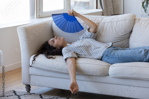 Exhausted young woman suffering from hot weather, overheat, too high air temperature at home, lying on sofa, waving paper handheld fan, cooling, feeling discomfort photo