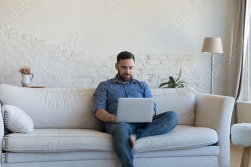 Focused freelancer man using laptop, holding computer on lap, sitting on couch, typing, working. Relaxed millennial guy resting on sofa, watching movie, playing online game. Communication concept
