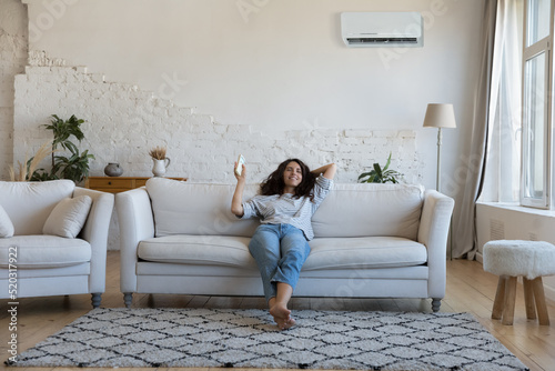 Happy pretty Latin woman enjoying cool conditioned air, relaxing on comfortable sofa, using remote control for turning AC on, starting cooling domestic equipment. Home appliance concept photo