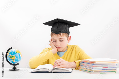 Schoolboy wears the students hat and does his homework. Bored or tired boy reads book sitting at desk. Portrait on white background.