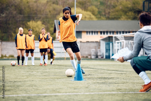Female soccer player leading ball among cones during sports training. © Drazen