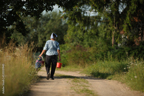 an old woman in a headscarf walks along a village road and carries heavy packages and a bucket