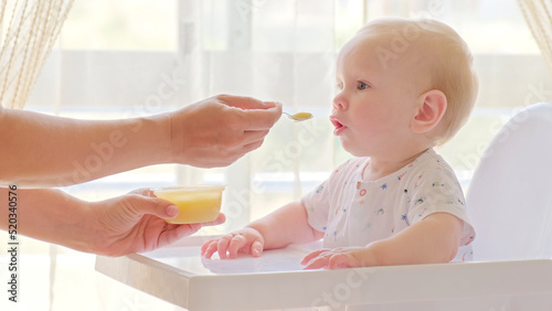 Mom feeds a 9 month baby with fruit puree from a spoon, close-up, high key. Mother is Feeding a baby from a spoon. Infant boy eats sitting on baby's chair. Mother cares about little son.