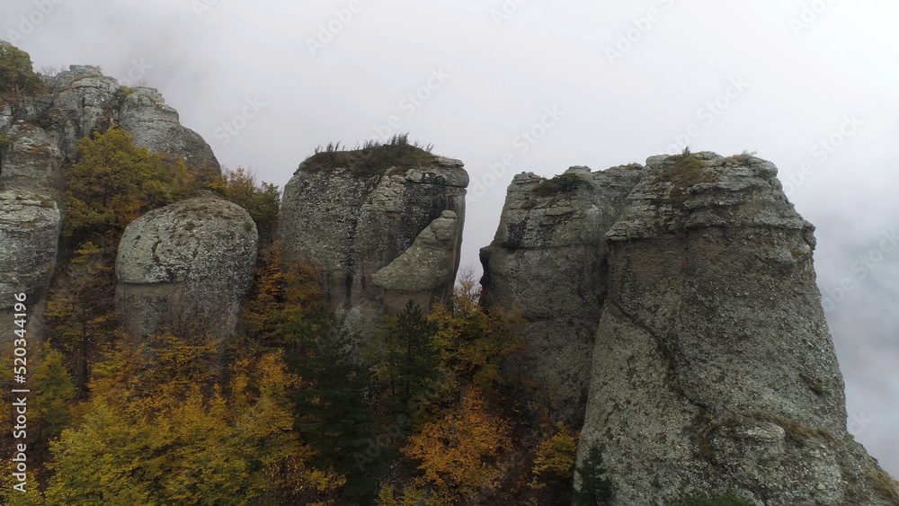 View near stone pillar of cliff. Shot. Top view of stone pillar of rock with approaching thick fog. Autumn landscape with colorful shrubs and dense gray fog on mountain