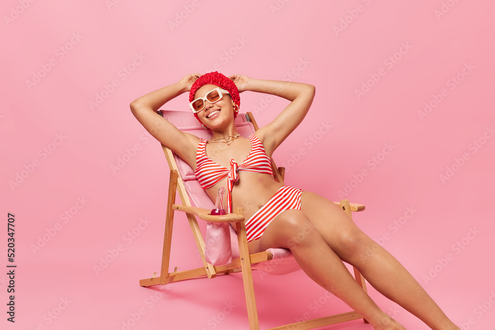 Happy relaxed young woman dressed in swimsuit bathhat and sunglasses poses on comfortable deck chair takes nap gets suntan enjoys summer vacation isolated over pink background. Perfect holiday - obrazy, fototapety, plakaty 