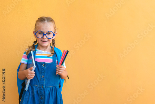 Little school girl in uniform and eyeglasses with backpack standing over yellow background, holding notebook and colored pencils, smiling
