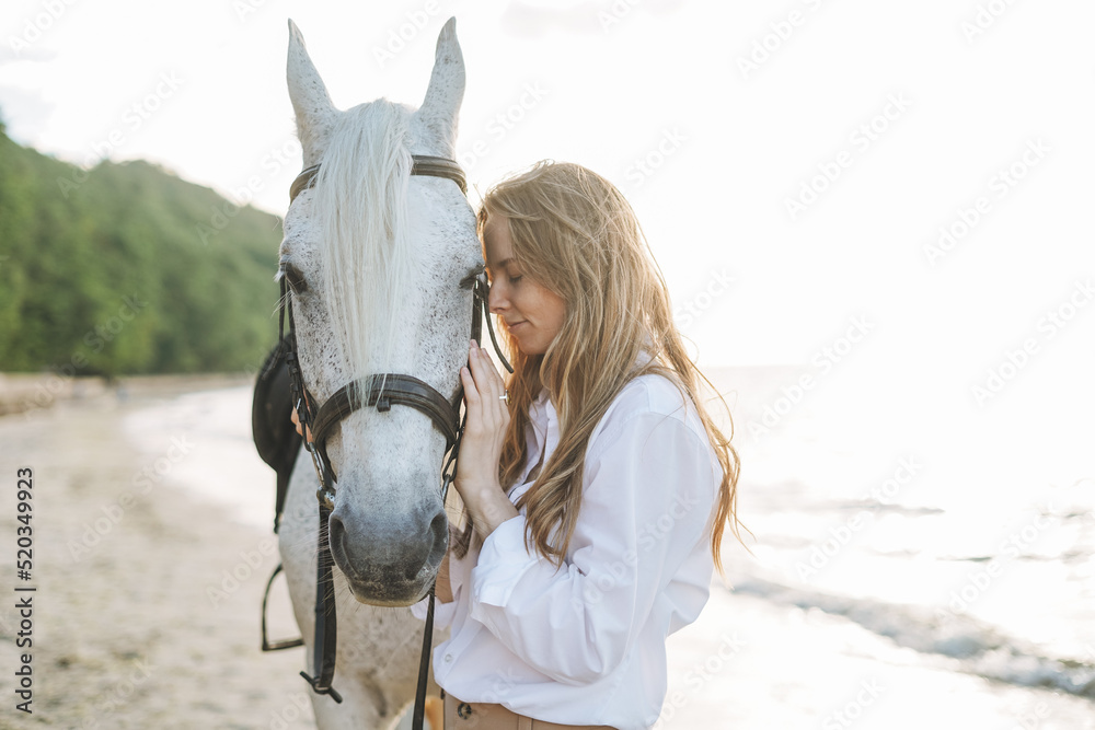 Obraz premium Young long hair woman in white shirt riding white horse on seascape background