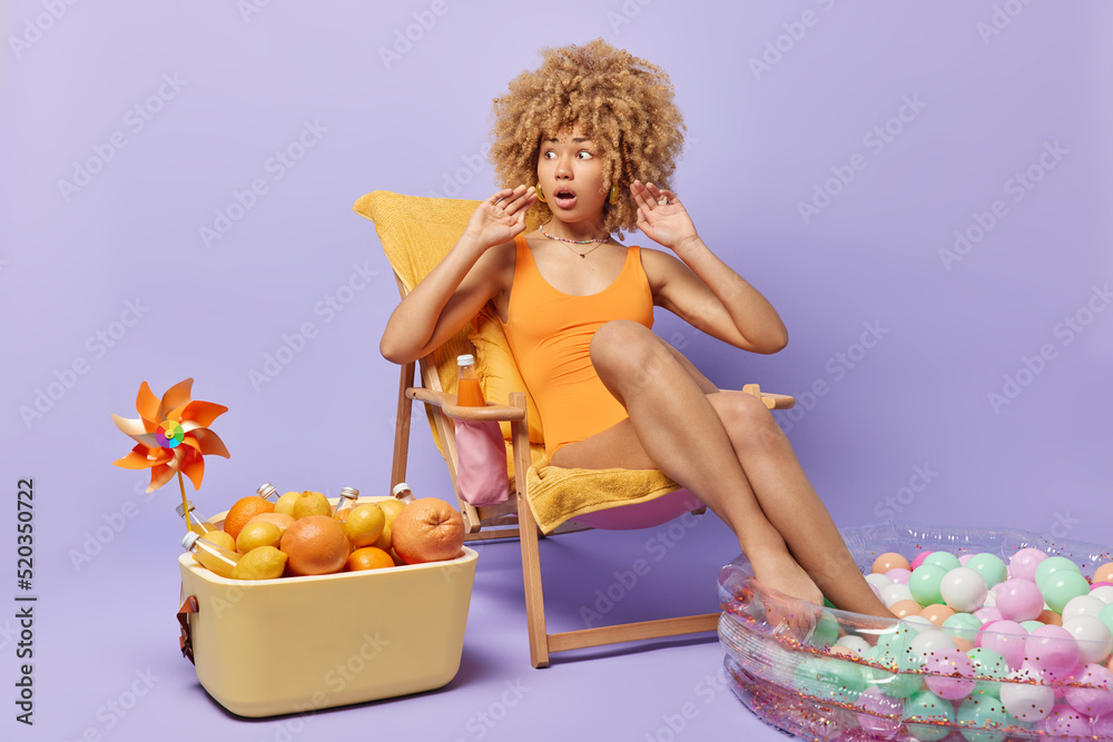 Beautiful amazed woman with curly hair reacts to shocking news dresed in swimsuit poses on comfortable deck chair keeps legs in inflated pool uses portable fridge isolated over purple background