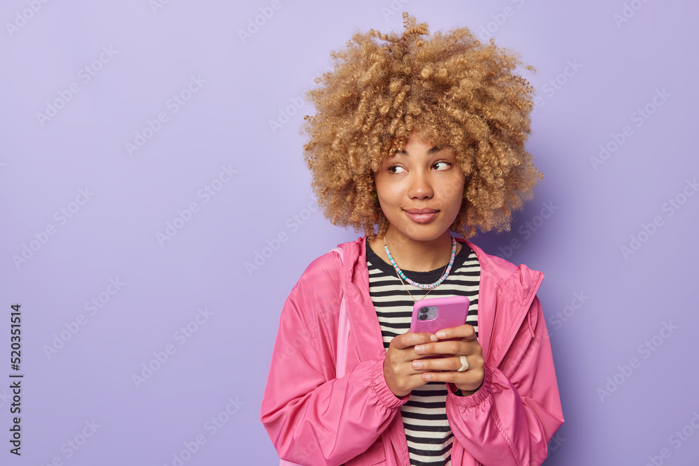 Studio shot of thoughtful dreamy young woman with curly hair looks away holds smartphone surfs through internet uses cellular application dressed casually isolated over purple studio background