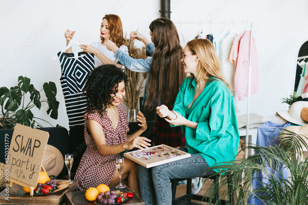 Fototapeta premium four young woman female caucasian and african students at swap party try on clothes, bags, shoes and accessories, change clothes with each other, second hand for things, zero waste life, eco-friendly