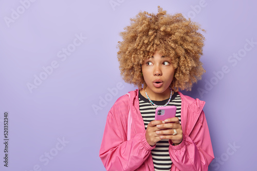 Surprised curly haired woman chats with friends or post in social networks holds mobile phone browses content looks amazed aside dressed in striped jumper and pink jacket isolated on purple wall