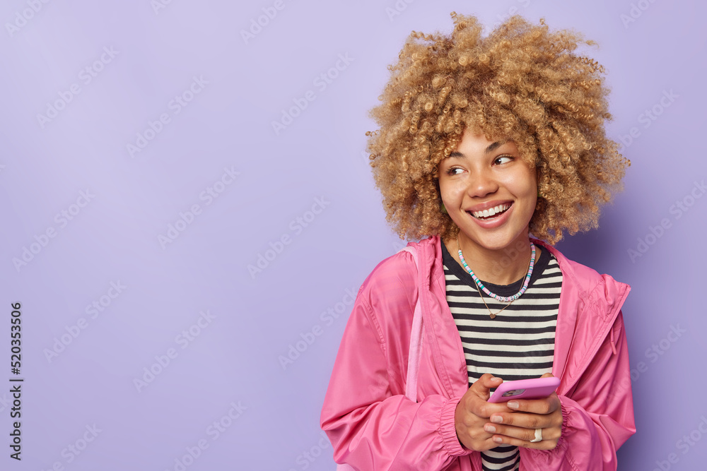 Happy beautiful woman with curly blonde hair sends mails via smartphone chats in social networks uses free internet connection for blogging looks away smiles broadly isolated over purple background