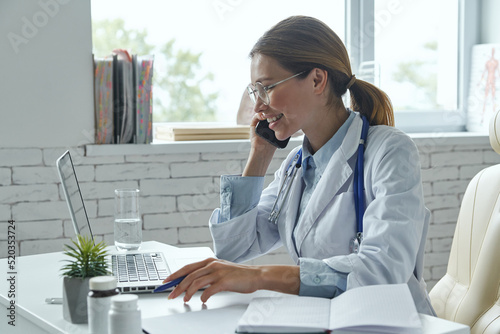 Cheerful female doctor talking on phone and using computer while sitting at the medical office