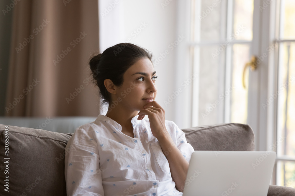 Dreamy beautiful young Indian woman holding laptop on lap, resting on couch, looking at window away, touching chin, thinking, d reaming, smiling at good thoughts, plans, ideas Dreamy beautiful young Indian woman holding laptop on lap, resting on couch, looking at window away, touching chin, thinking, d reaming, smiling at good thoughts, plans, ideas