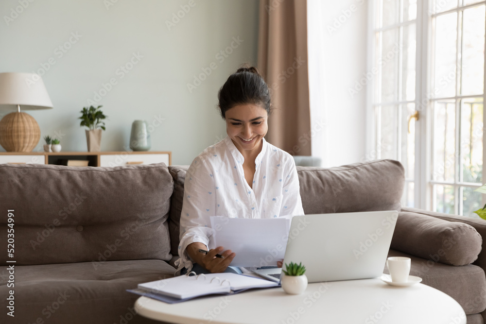 Happy Indian freelance employee woman reading paper document at laptop, smiling at good news, sales report, job result, contract, deal conditions, sitting on couch at laptop, working at home Happy Indian freelance employee woman reading paper document at laptop, smiling at good news, sales report, job result, contract, deal conditions, sitting on couch at laptop, working at home