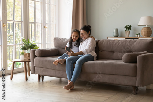 Happy Indian mom and kid taking home selfie on mobile phone, resting on sofa in living room interior, laughing, hugging, holding smartphone in outstretched hand, making video call. Full length