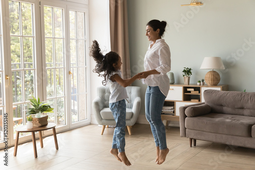Excited Indian kid and happy mom holding hands, jumping to music on floor. Cheerful mother teaching kid to dance music in living room, smiling, laughing, having fun. Family activity concept