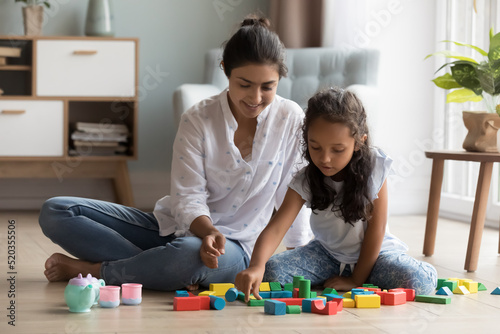 Cheerful pretty Indian mother and little daughter kid playing on heating floor at home, arranging colorful toy blocks. Nanny, daycare teacher entertaining child, enjoying learning game