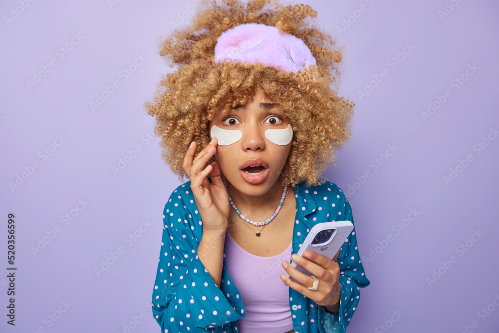 Amazed puzzled woman applies white beauty patches under eyes to reduce wrinkles undergoes skin care procedures before sleep holds cellular dressed in comfortable pajama isolated over purple wall Amazed puzzled woman applies white beauty patches under eyes to reduce wrinkles undergoes skin care procedures before sleep holds cellular dressed in comfortable pajama isolated over purple wall