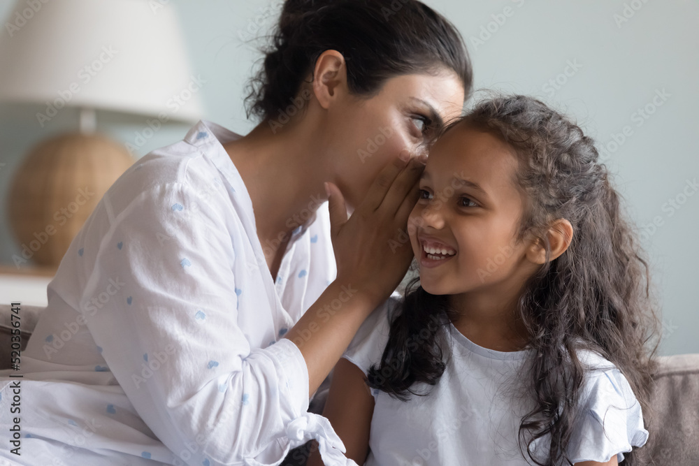 Young Indian mom sharing secret with happy daughter, whispering in ear. Excited little kid looking at away, laughing, listening to mother. Mum and child enjoying friendship, trust, having fun together Young Indian mom sharing secret with happy daughter, whispering in ear. Excited little kid looking at away, laughing, listening to mother. Mum and child enjoying friendship, trust, having fun together