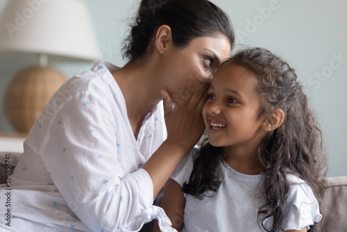 Young Indian mom sharing secret with happy daughter  whispering in ear. Excited little kid looking at away  laughing  listening to mother. Mum and child enjoying friendship  trust  having fun together