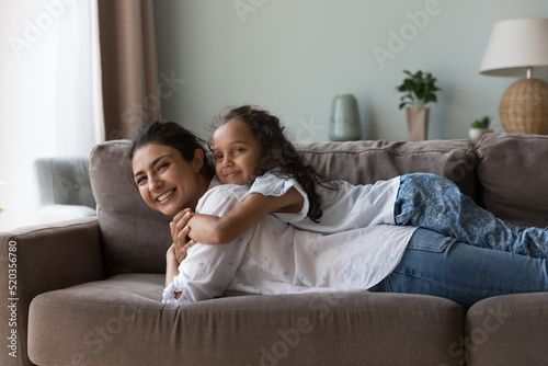 Happy sweet Indian kid girl resting on happy moms back, smiling. Cheerful pretty young mother lying on sofa at home, piggybacking daughter child, enjoying leisure, entertainment, playing active games