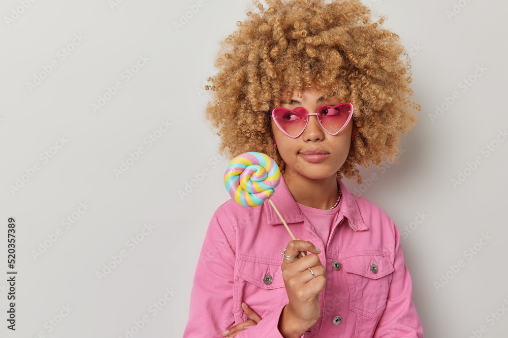 Studio shot of thoughtful young woman with curly hair focused away holds delicious caramel candy on stick wears sunglasses and jacket poses against grey background blank space for your promotion Studio shot of thoughtful young woman with curly hair focused away holds delicious caramel candy on stick wears sunglasses and jacket poses against grey background blank space for your promotion