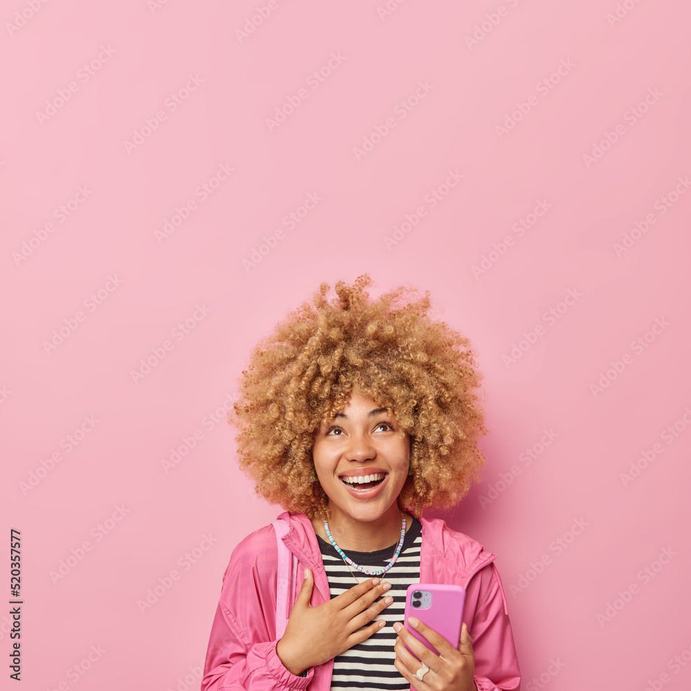 Vertical shot of positive woman with curly bushy hair keeps hand on chest smiles gladfully dressed in striped jumper and jacket holds mobile phone hears something funny isolated over pink wall