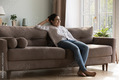 Cheerful carefree young Indian woman enjoying relaxation co comfortable sofa, looking at window away, dreaming, thinking over good news, plans. Home leisure concept. Full length