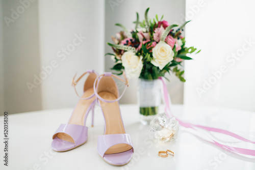 Lilac bride's sandals on the table with the bride and groom's wedding rings and a bouquet of flowers in a vase.