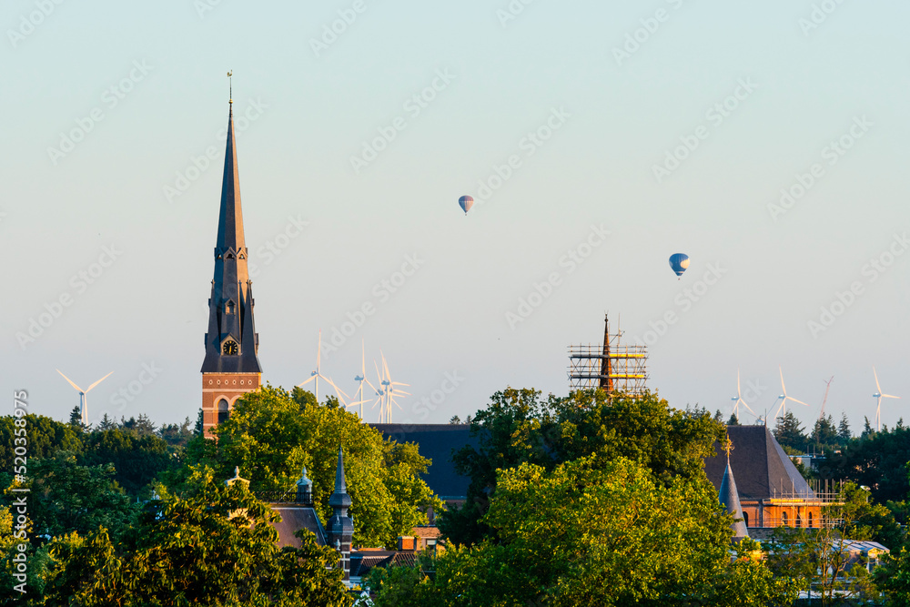 Fototapeta premium European cathedral tower surrounded by green trees at sunset with hot air balloons. Breda, the netherlands