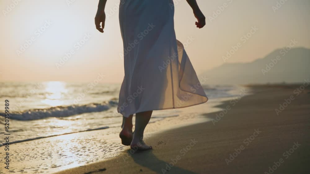 Barefoot woman in a summer dress walking along the seashore at sunset Barefoot woman in a summer dress walking along the seashore at sunset