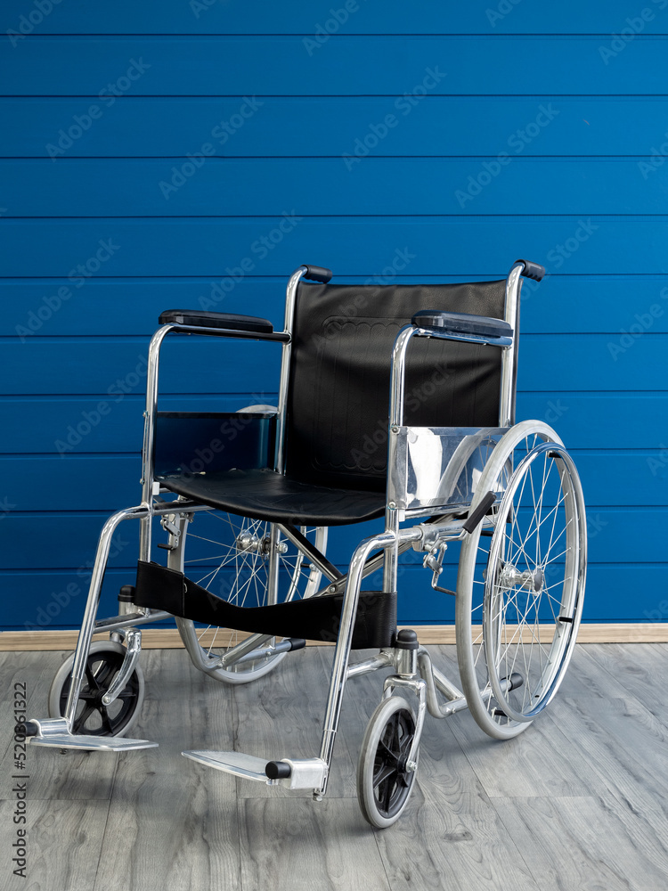Empty steel wheelchair with black leather seat and backrest on the blue wooden plank wall background, vertical style. Chair with wheels for use as transport by person who unable walk, illness, injury.