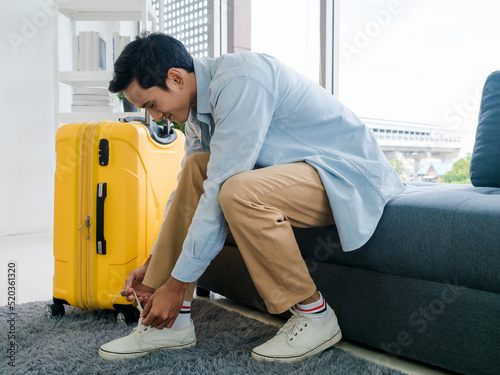 A man with yellow luggage. A male in denim shirt and brown trousers sitting on sofa tying shoelaces near the suitcase in office room. Summer holiday travel vacation concept. Time to journey.