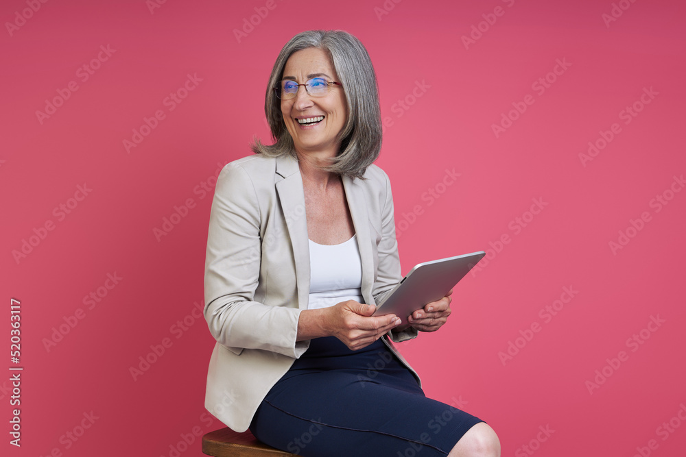 Happy senior woman in formalwear looking away while using digital tablet against pink background Happy senior woman in formalwear looking away while using digital tablet against pink background