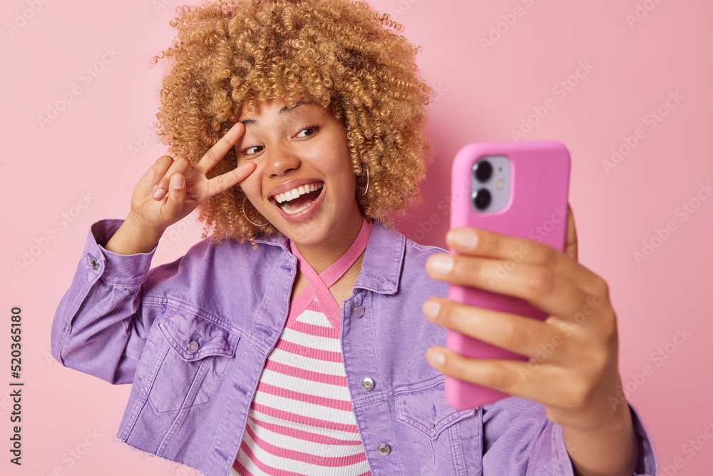 Glad smiling woman with curly hair smiles broadly makes peace gesture over eye poses for selfie in smartphone camera sends positive vibes dressed in purple jacket isolated on pink background