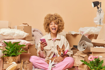 Happy curly haired woman dressed in casual clothes sits in lotus pose holds mobile phone plays with pet focused overhead prepares for relocation surrounded by carton boxes isolated on beige background