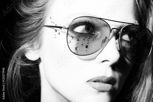 Fashion and make-up concept. Beautiful girl with retro sunglasses and long hair close-up studio portrait. Model with glued jewelers around her eyes looking aside the camera. Black and white image