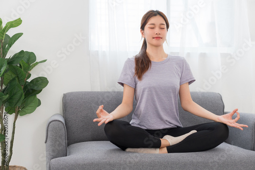 Yoga exercise concept, Young Asian woman doing yoga exercise in lotus pose on couch in living room