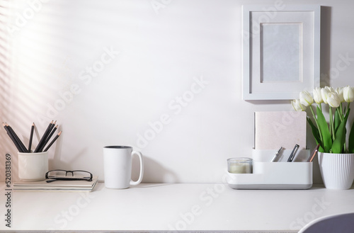 Coffee cup, stationery and flower pot on white table with sunlight through blinds by the window.