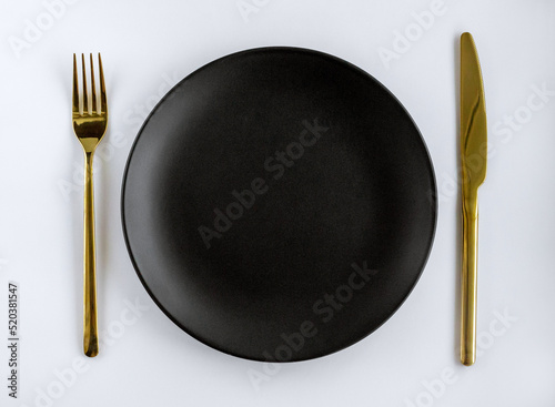Black empty ceramic plate with golden cutlery on a white background. Center composition.
