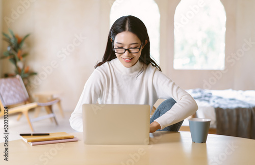 Beautiful smiling freelancer Asian woman in glasses sitting workplace with laptop in home winter atmosphere. Beautiful stylish Japanese lady Millennial enjoying working at home. New Normal Concept. © edward_indy