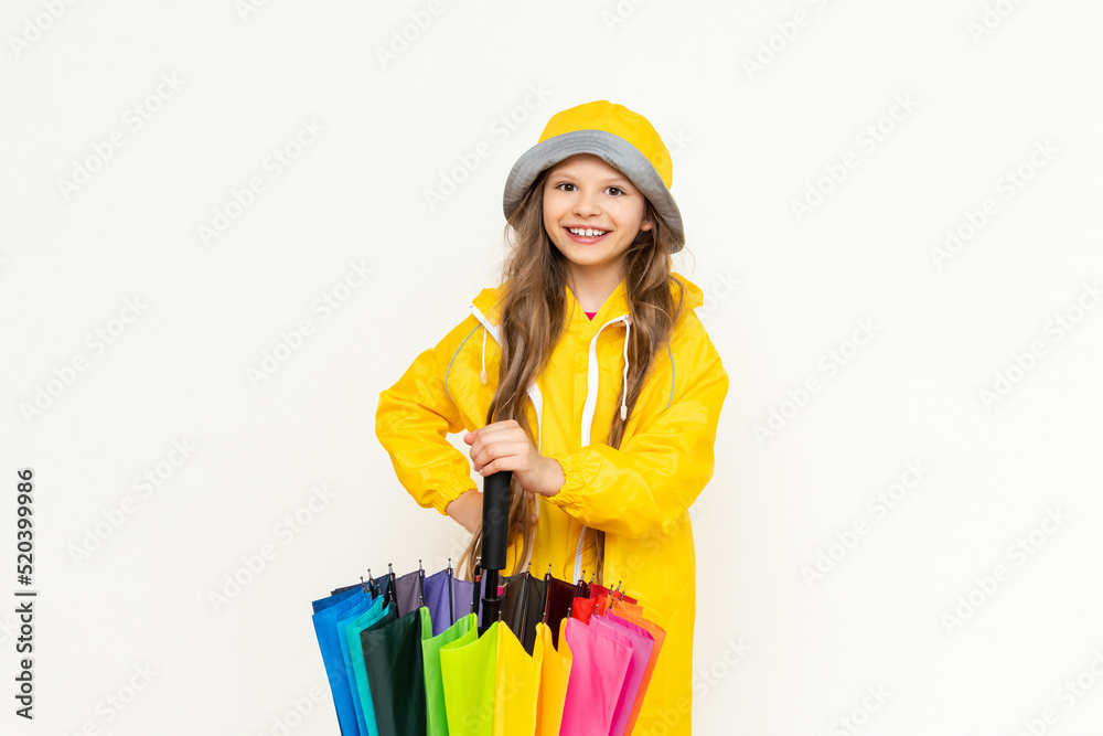 A little girl in a yellow raincoat and panama hat on a white isolated background leans on a multicolored rainbow umbrella. A little girl in a yellow raincoat and panama hat on a white isolated background leans on a multicolored rainbow umbrella.