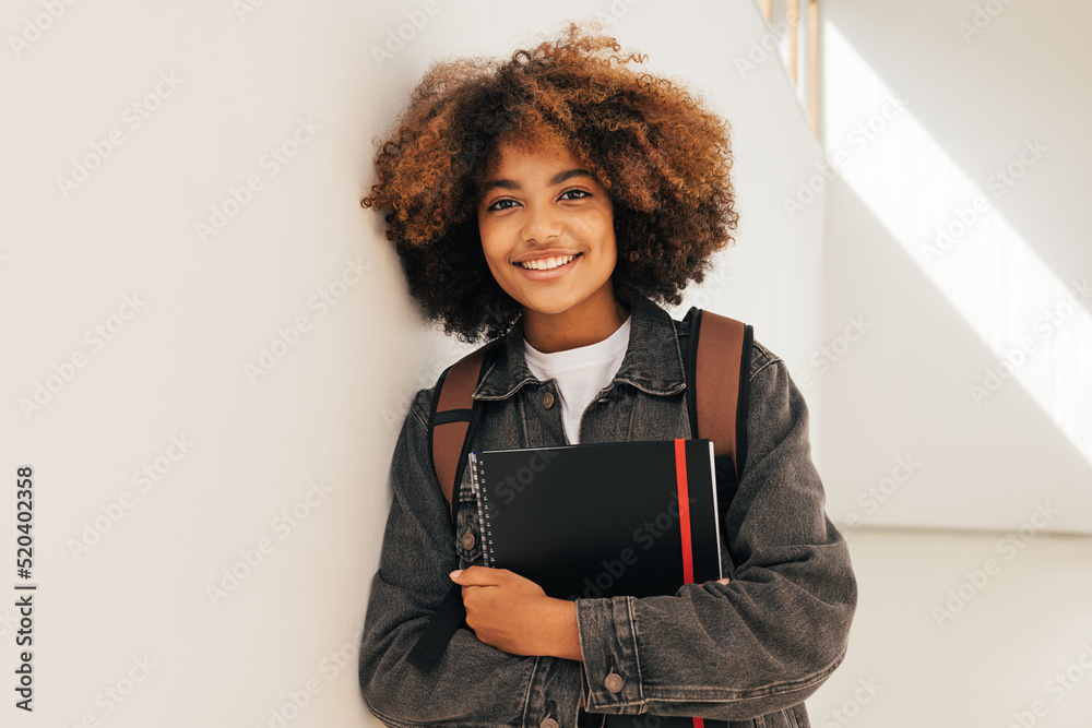 Portrait of a young beautiful female student leaning a wall. Smiling girl looking at camera while standing in high school. - obrazy, fototapety, plakaty 