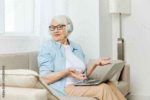 a happy modern old lady looks at the monitor holding a laptop on her lap talking through headphones smiling happily and gesticulating with her hands © Tatiana