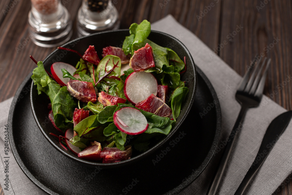 In a deep black plate, a mix of lettuce leaves with radishes and red orange slices, seasoned with olive oil, Black cutlery. In a deep black plate, a mix of lettuce leaves with radishes and red orange slices, seasoned with olive oil, Black cutlery.